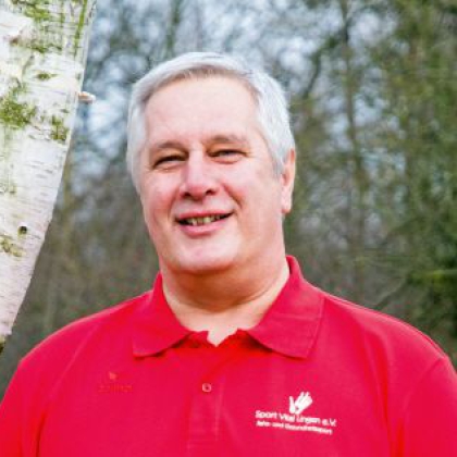 Ein lächelnder Mann in einem roten Poloshirt steht draußen vor einem Birkenbaum und einem grünen Waldhintergrund.